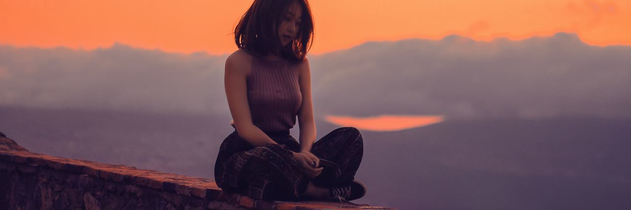 young woman sitting on wall at sunset in yoga meditation pose