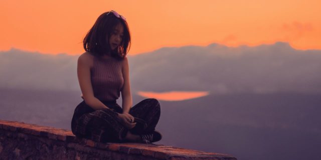 young woman sitting on wall at sunset in yoga meditation pose