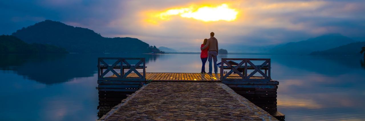 photo of couple standing on pier over water at sunset