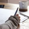 close up photo of woman's hand holding pen and writing with coffee mug in background