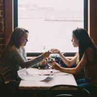 photo of two women eating together in front of window and brick wall