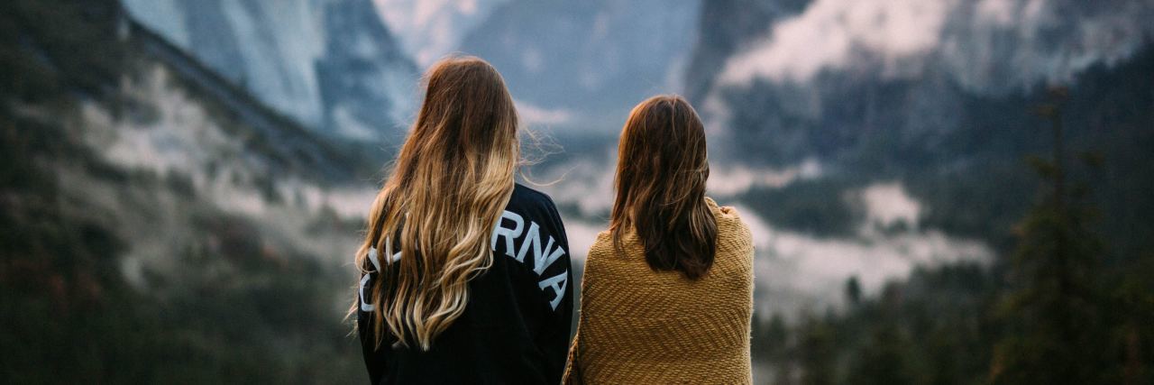 photo of two women standing in front of mountain range and forest together looking out over scenery