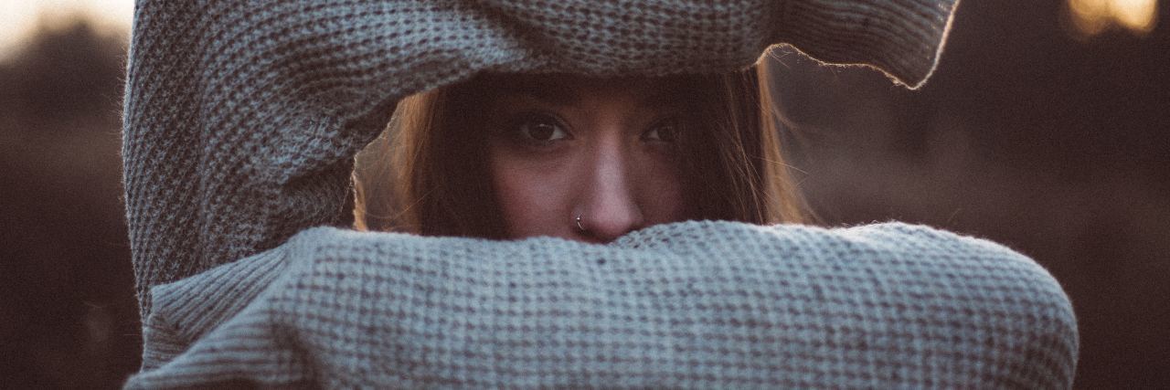 How People React to My Self-Harm Scars at the Gym photo of woman holding up arms in front of her face and looking through the gap. Her arms are horizontal and she's wearing a sweater.