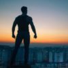 photo of man silhouetted against sunset sky in superhero pose, fists clenched and confident, looking out over city