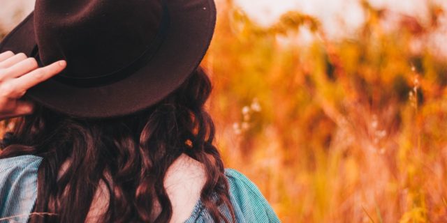 photo of woman wearing hat over long brown hair standing in front of orange grass or wheat