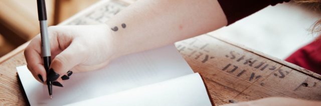 close up photo of woman writing in notebook with semicolon tattoo on wrist