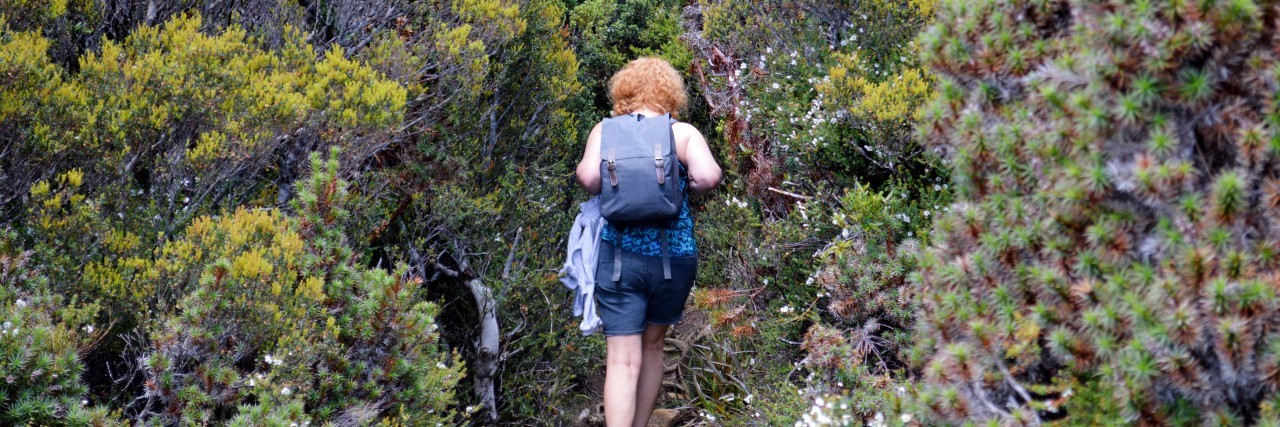 photo of woman walking along dirt trail with a blue backpack and shrubbery on either side