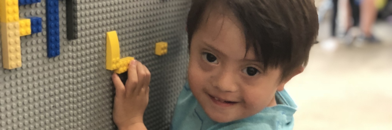 Boy with Down syndrome looking at camera while holding on to lego pieces on a lego board wall.