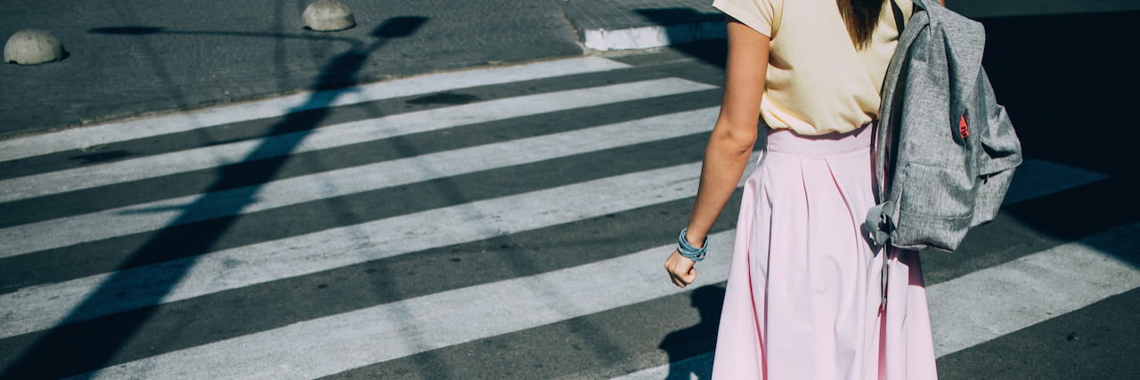 Rear view of young woman wearing pink skirt with backpack crossing the road at pedestrian crosswalk on sunny summer day.