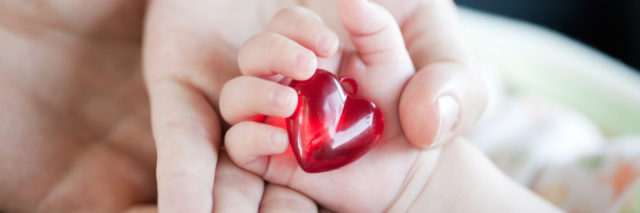 A woman holding a baby's hand. The baby holds a plastic red heart.