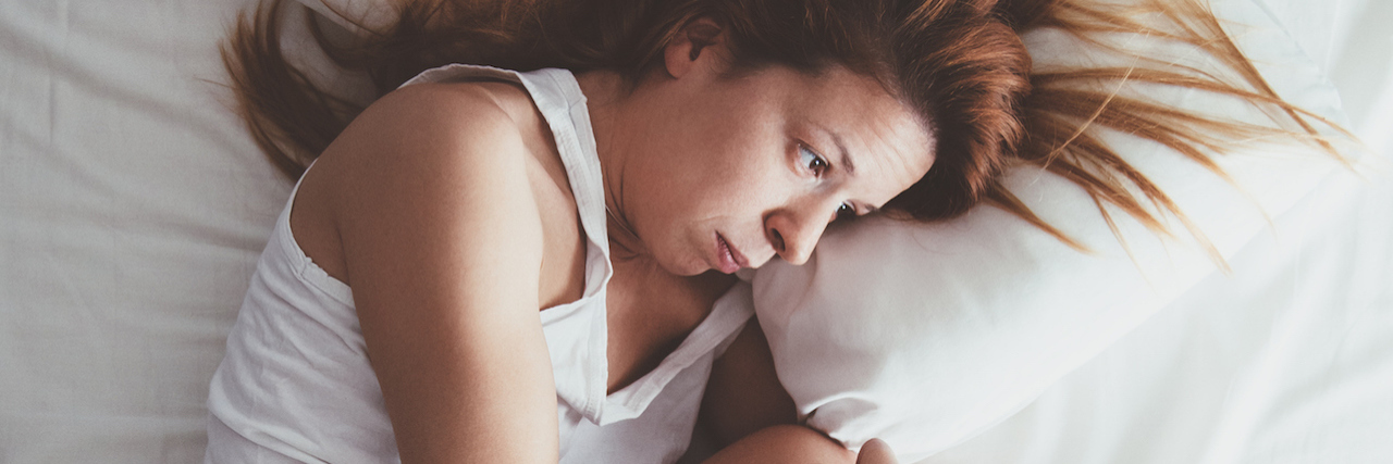 A woman lying in bed in a fetal position, wearing all white, her hair flowing around her head