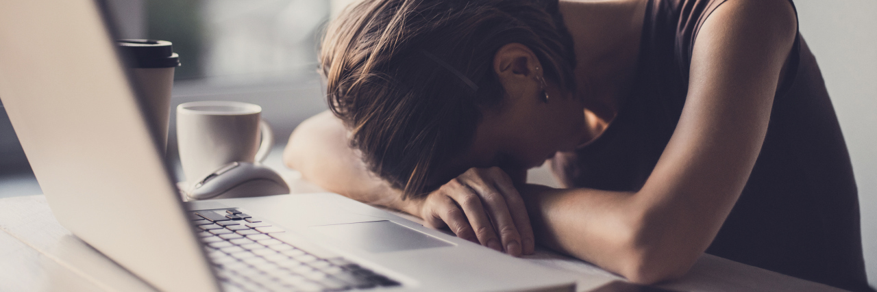 Reasons Office Life Is Hard When You Have a Chronic Illness photo of office worker woman resting head on arms in office in front of laptop