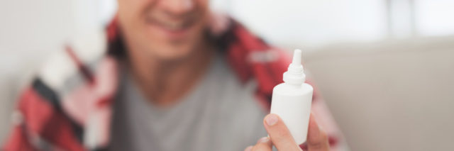A man with a cold sits on the couch, hiding behind a red rug. He sprinkles a special nasal spray into his nose. The man is very satisfied with the spray