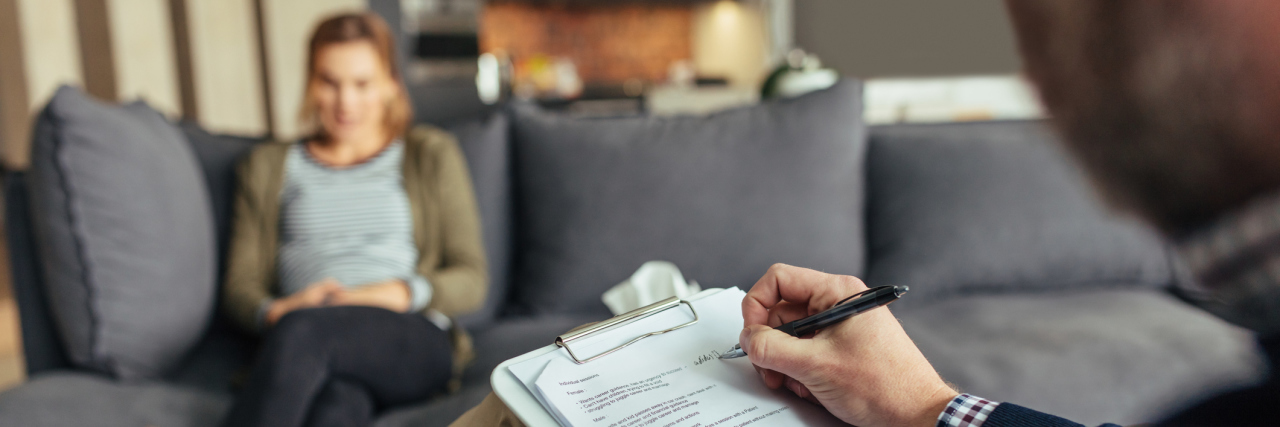 My Therapist Thinks I Hate Myself Psychologist taking down notes during therapy session with female patient. Psychotherapist with clipboard and woman patient sitting in background.