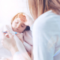 Worried mother putting her right hand on the forehead of her daughter and keeping thermometer in left hand while sitting next to her kid