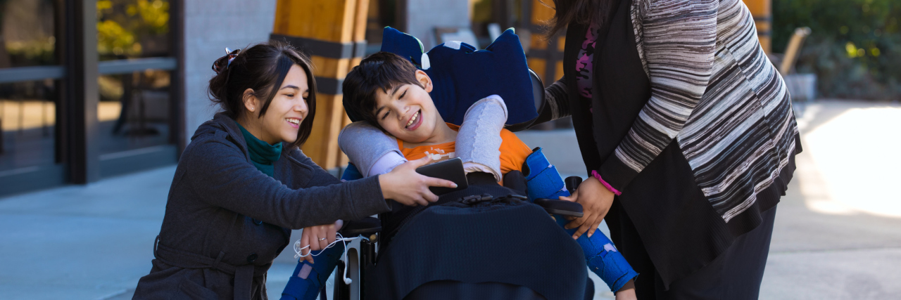 Boy with a disability in a wheelchair watching a video on one of his two caregiver's smartphone.