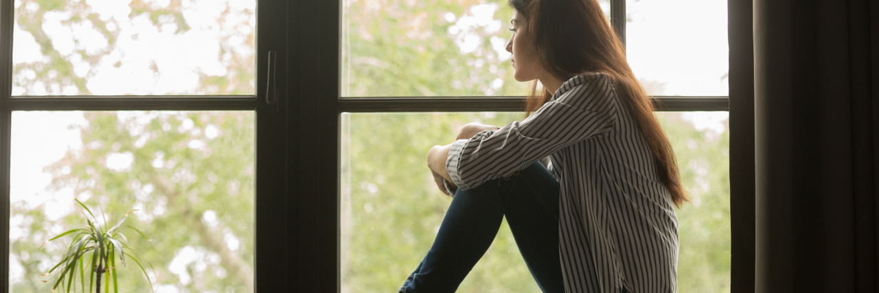 photo of woman sitting on windowsill looking out of window thoughtfully and hugging her knees