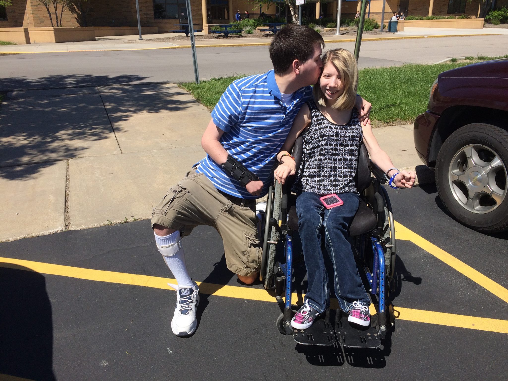 Sarah and Joe in a parking lot. Sarah sits in a wheelchair while Joe is on one knee beside her, kissing her cheek. He wears a leg brace.