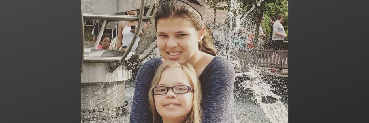 How My Daughter Has Been Impacted by Her Sister With Down Syndrome Big sister hugs little sister resting her chin on sister's head
