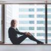photo of young woman sitting on window still looking out of large windows with wistful look