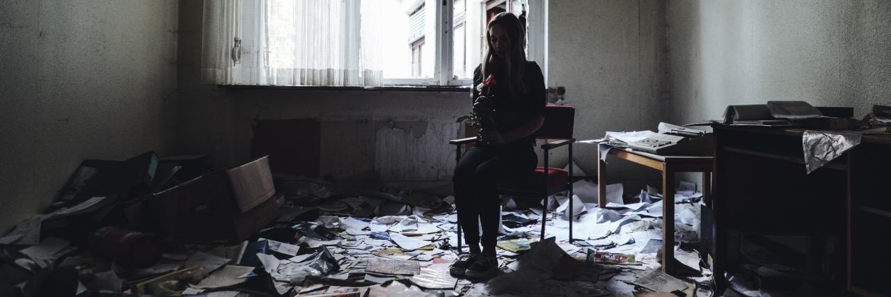 Burying Your Trauma in Mental 'Storage Boxes' photo of woman holding rose and sitting on hair in abandoned room with papers all around floor