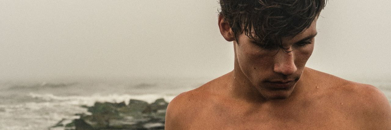 photo of man without shirt standing on misty rocky beach looking away from camera