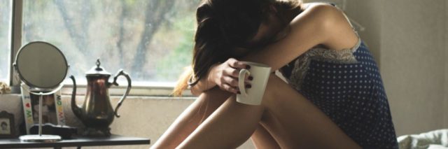 photo of woman looking tired sitting on bed with head resting on knees and cup or mug in hand