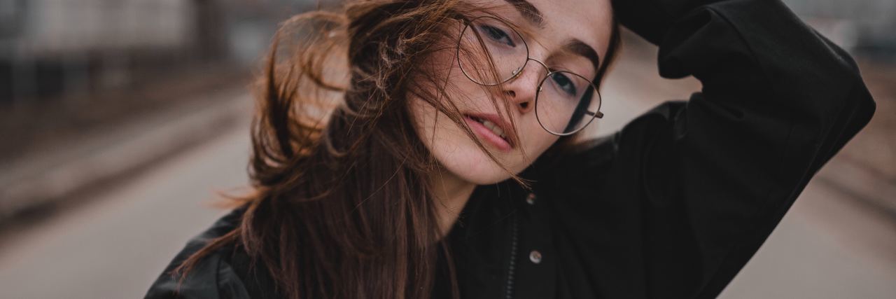 Feeling Ashamed Because You Seem Better in Your Mental Illness photo of young woman wearing glasses standing in front of camera in heavy coat with hair in wind and hand in hair