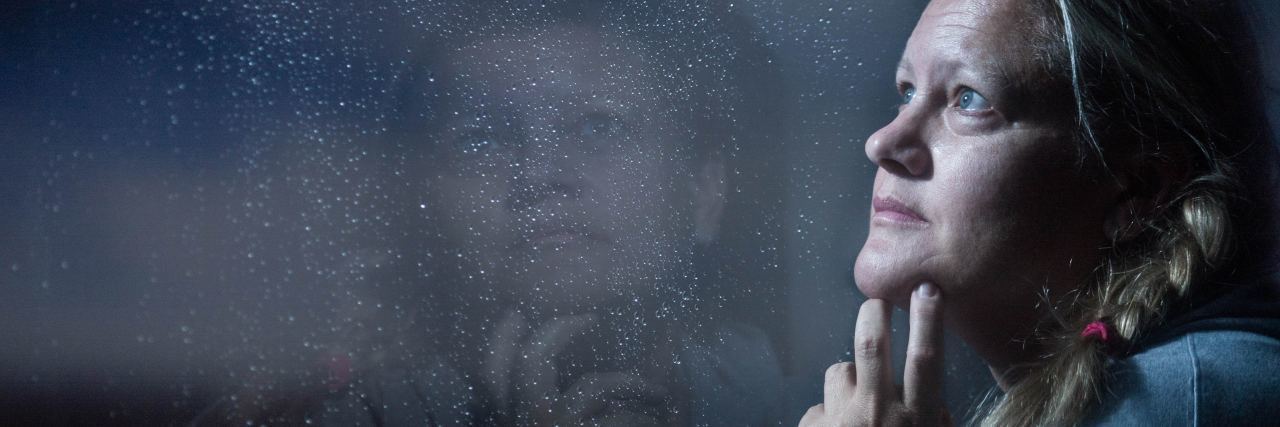 photo of middle-aged woman looking thoughtfully into dark window with reflection