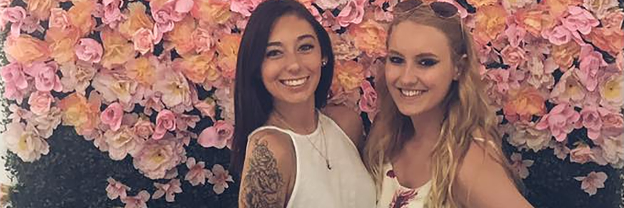 To the Best Friend With the Same Chronic Pain as Me photo of contributor with her best friend in front of wall with flower petals and words saying skinny dip. they are both smiling for camera and posing.