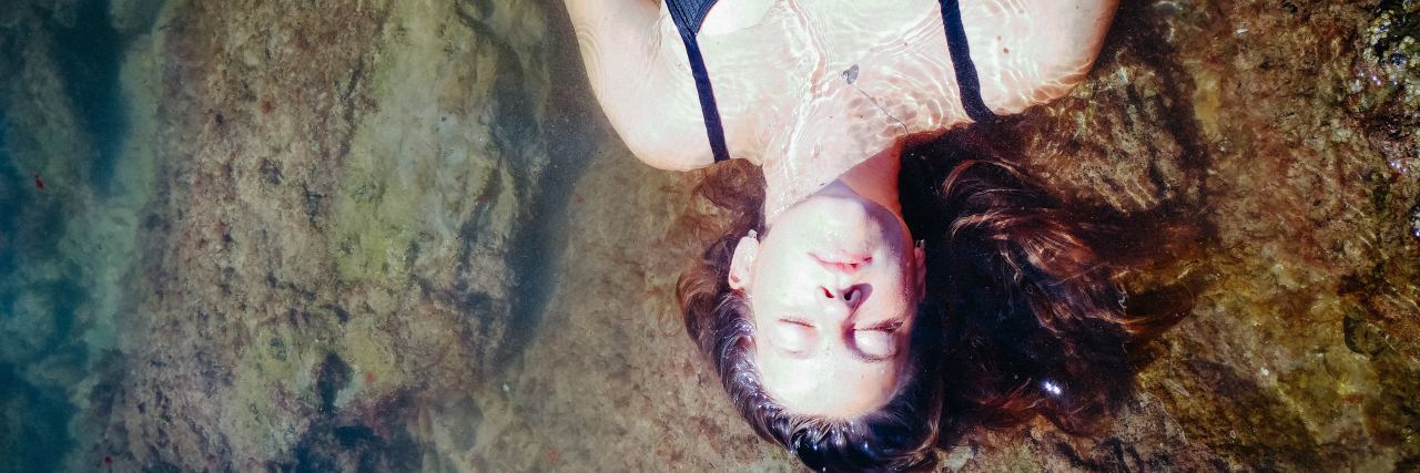 Don't Ask Someone With a Chronic Illness 'Are You Better?' A woman lying on a rock in the water, her eyes closed