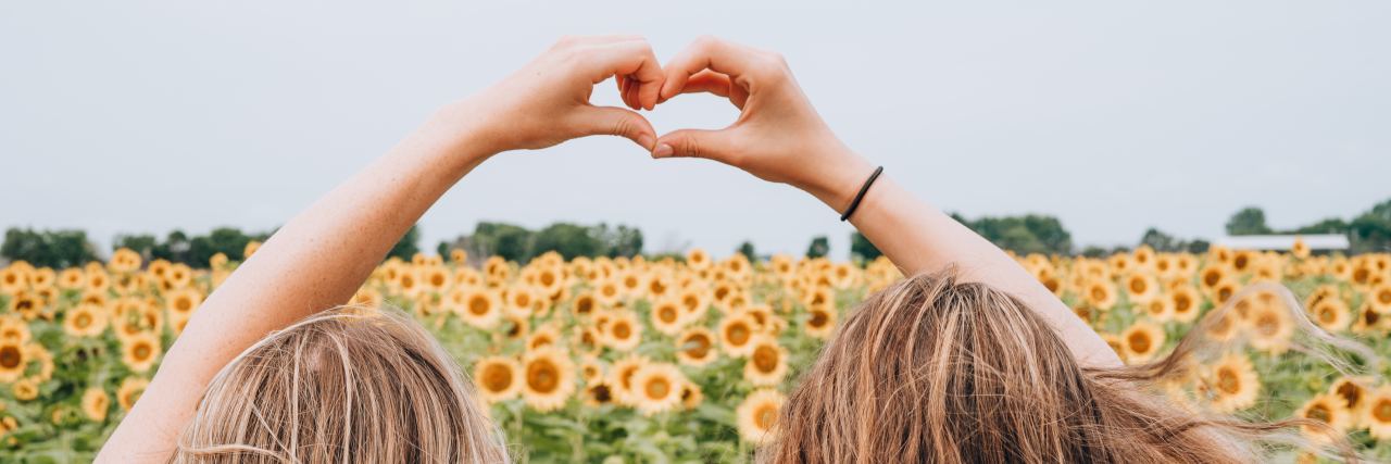 photo of two women with blonde hair standing in front of sunflowers and making heart sign with joined hands