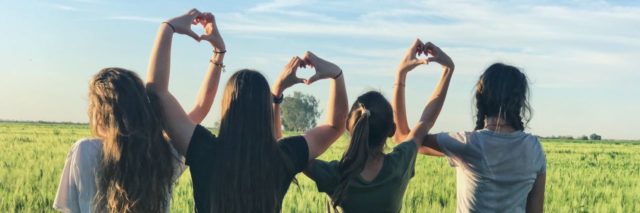 photo of four women standing in green wheat field in sunlight with arms linked, making heart symbol with each others' hands
