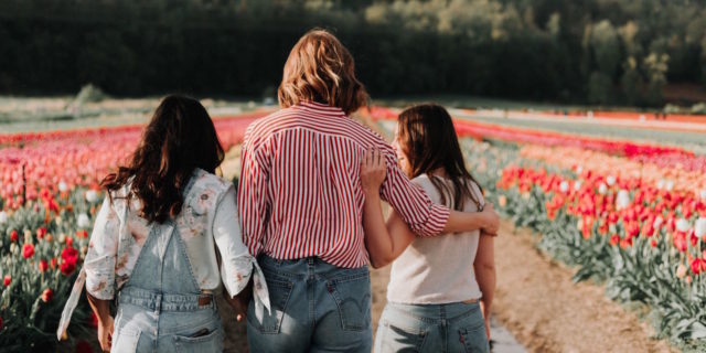 A group of friends facing away, arms around each other. They're standing in a field of flowers.