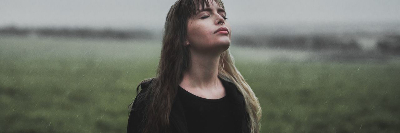 photo of woman standing in field on cloudy day with eyes closed and head turned to sky