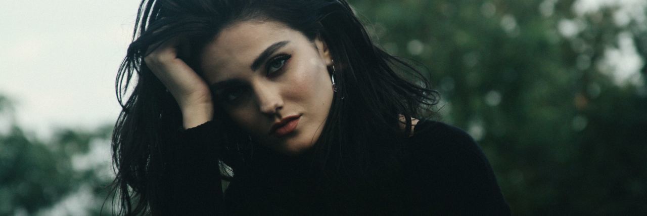 photo of young woman resting head on hand against backdrop of trees looking into camera with worried expression