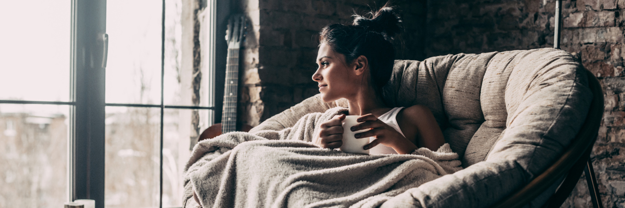 Why It's OK to Take Time for Yourself If You Can't Always Give 100% Attractive young woman covered with blanket looking through window and holding a cup while sitting in an armchair at home