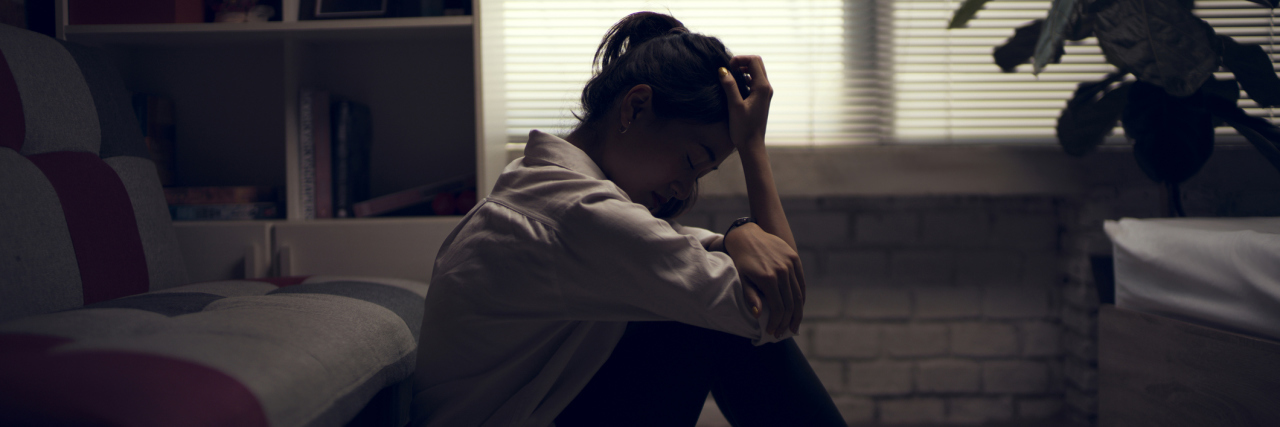 When Chronic Illness Makes You Feel Trapped and Unsafe in Your Body woman sitting on the floor in her living room. her head is in her hands and she appears upset. the blinds are drawn so the room is dark.