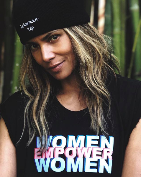 Halle Berry wearing a black beanie that says "woman up" and a black shirt that says "women empower women" in blue, pink and white lettering