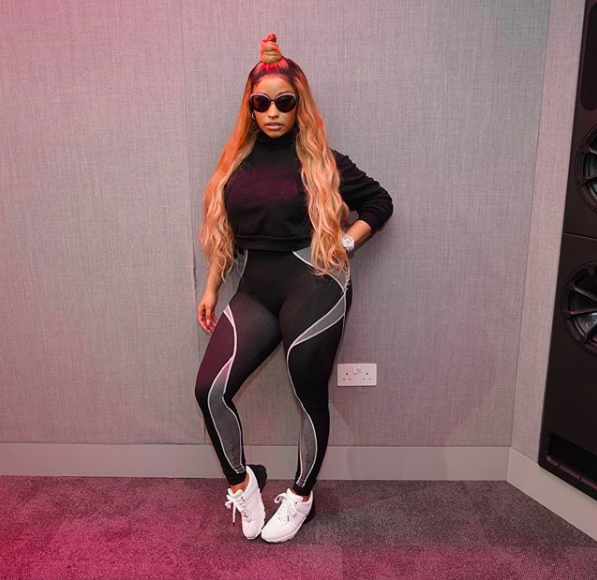 Nicki Minaj standing against a wall, wearing a black shirt, black and light gray pants, and white and black shoes.