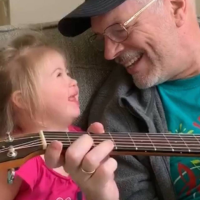 Grandpa playing guitar and looking at his granddaughter who looks up at him with a smile.