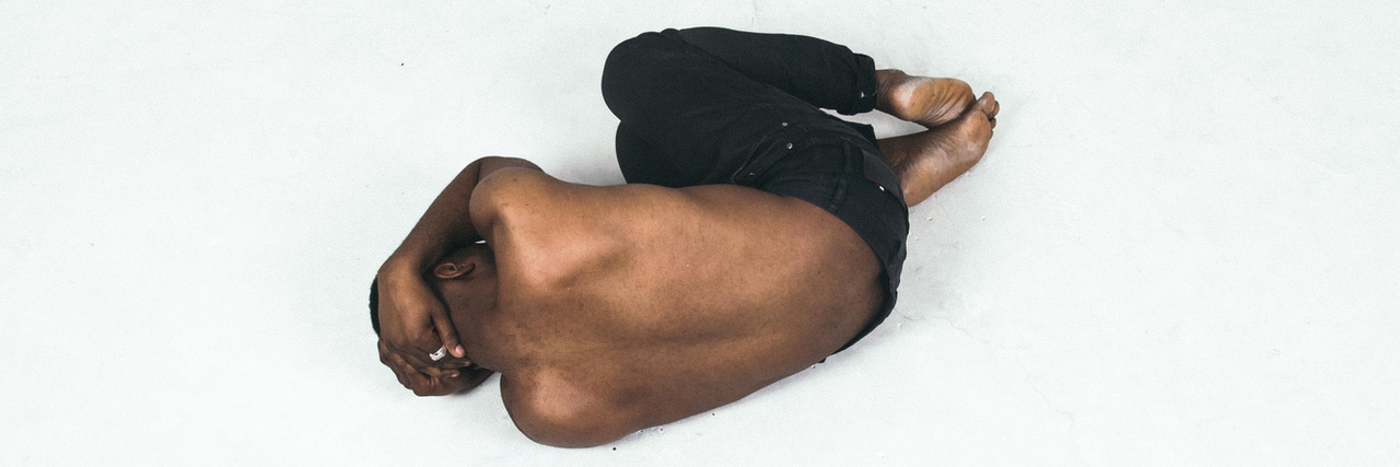 A black shirtless man laying in fetal position on the floor with his hands covering his head.