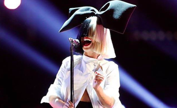 Sia singing into a microphone, wearing a wig covering her face with a oversize bow on top