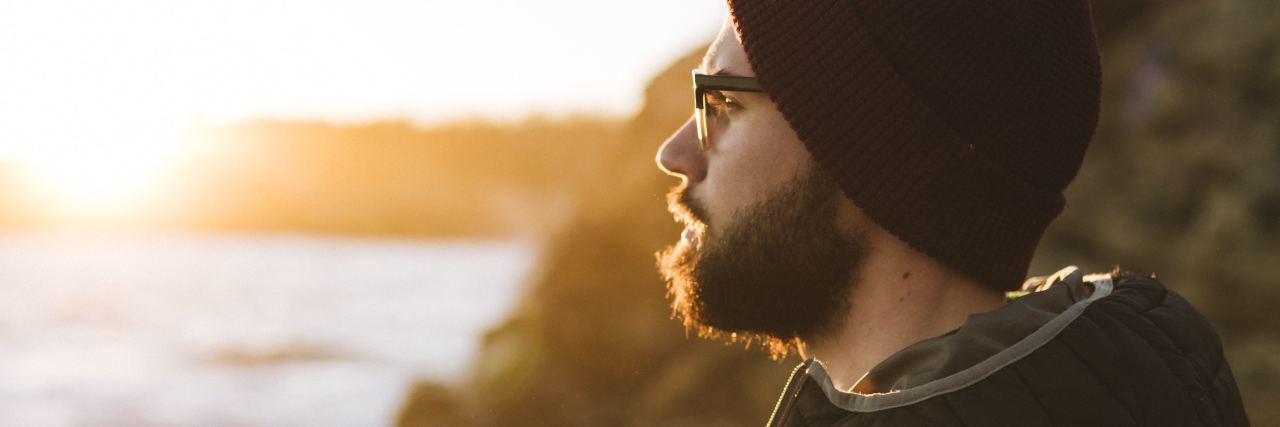 photo of man with beard wearing coat and hat and looking out over sea at sunset or sunrise