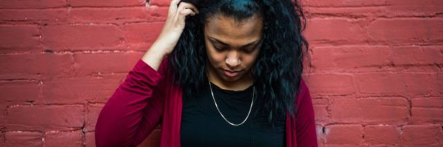 photo of black woman with hand in hair standing in front of red brick wall and looking down