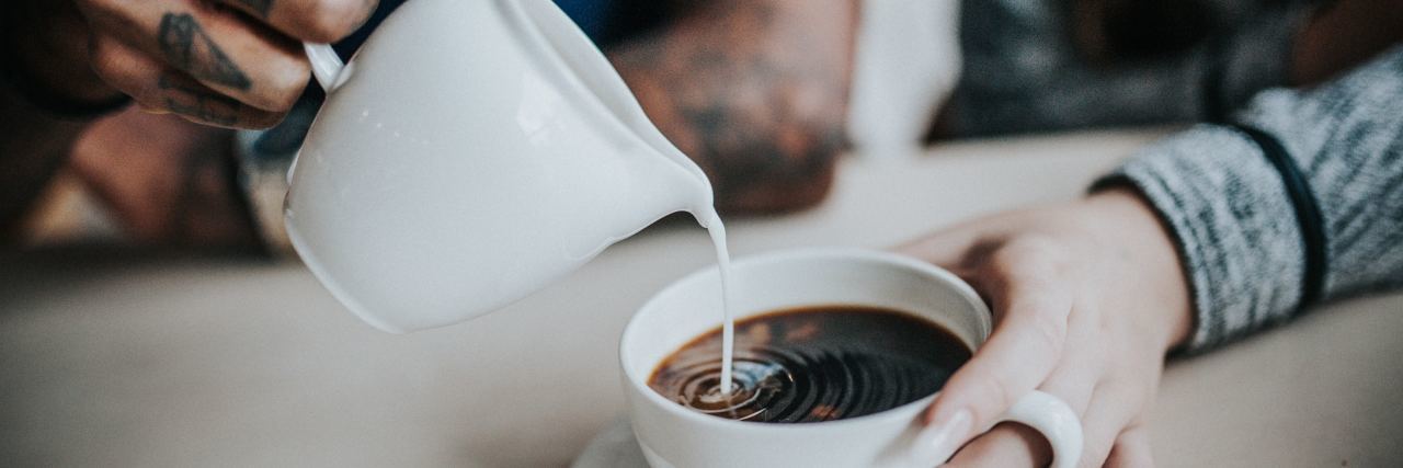 photo of woman holding coffee mug while man with hand tattoos pours cream or milk