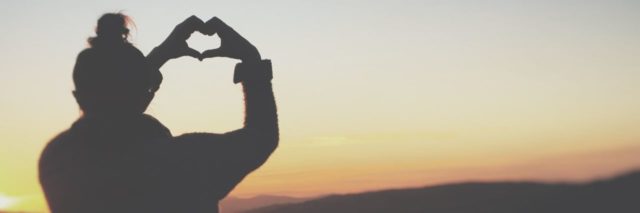 photo of woman making heart shape with hands silhouetted against sunset