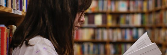 photo of young high school student with dark hair and glasses reading book alone in library
