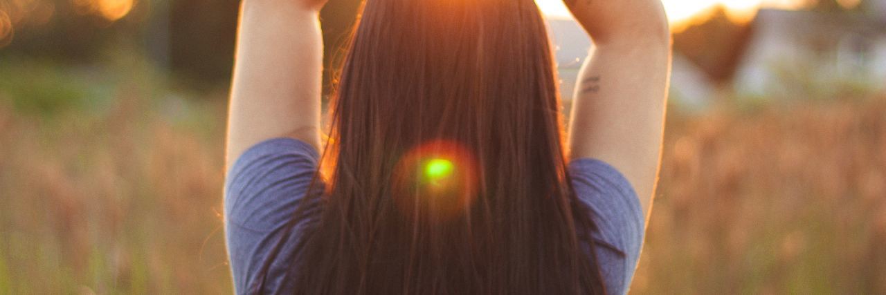 Don't Ask a Person With Chronic Illness How They Spend Their Time photo of woman standing in front of sunset or sunrise with hands raised and arms crossed above her head