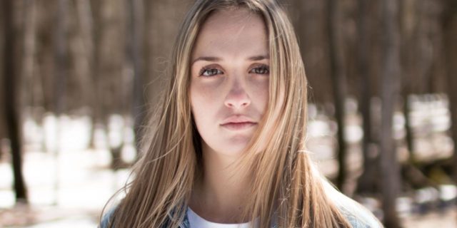 photo of young blonde woman standing in forest with trees behind her